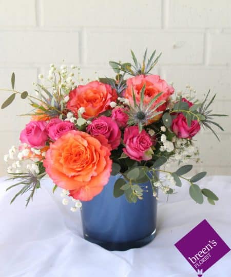 A classic new color combination is hot pink and orange. We created this arrangement using those bold colors in honor of Grace Goad’s “Hot Pink Orange Teal.” The two colors complement, yet contrast each other in this deep blue cylinder with touches of blue thistle and some baby’s breath to give these colors even more contrast.