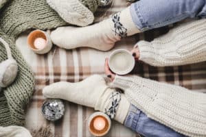 Young woman sits on plaid in cozy knitted woolen white sweater and socks holds cup of cocoa in her hands. Hygge New Year, cozy Christmas, preparation for holidays. Candles, Christmas balls.