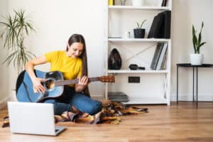 Hobbies and leisure activities during quarantine. Online training, online classes. A young woman watches a video lesson on playing the guitar, she sits on a cozy plaid with a guitar